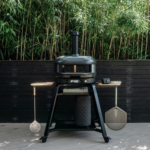 Gozney Dome Pizzaofen auf Dome & Dome XL Gestell mit Holzablagen und aufgehängtem Pizza-Zubehör auf einer Terrasse vor Bambushecke