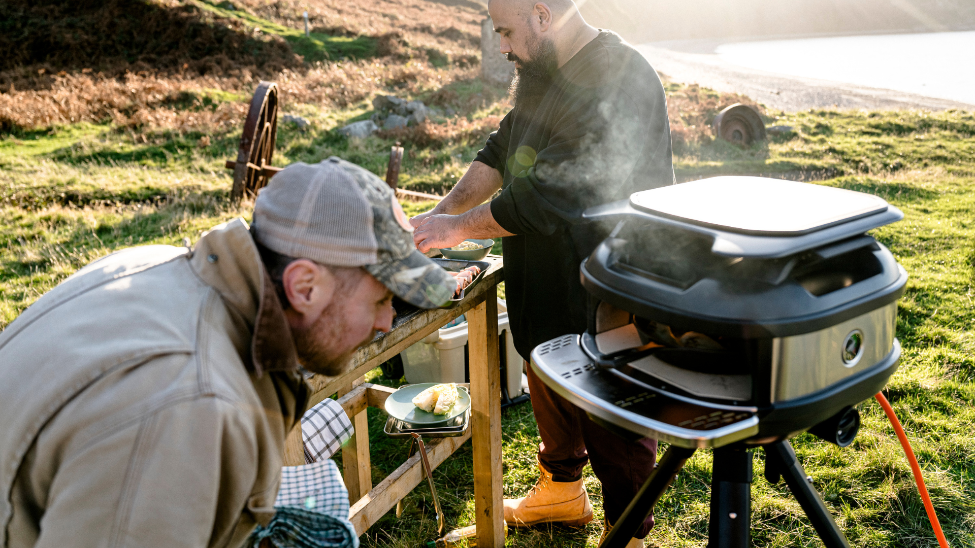 Zwei Personen bereiten Speisen am Gozney Tread Pizzaofen in Schwarz beim Outdoor-Kochen auf einer Wiese zu
